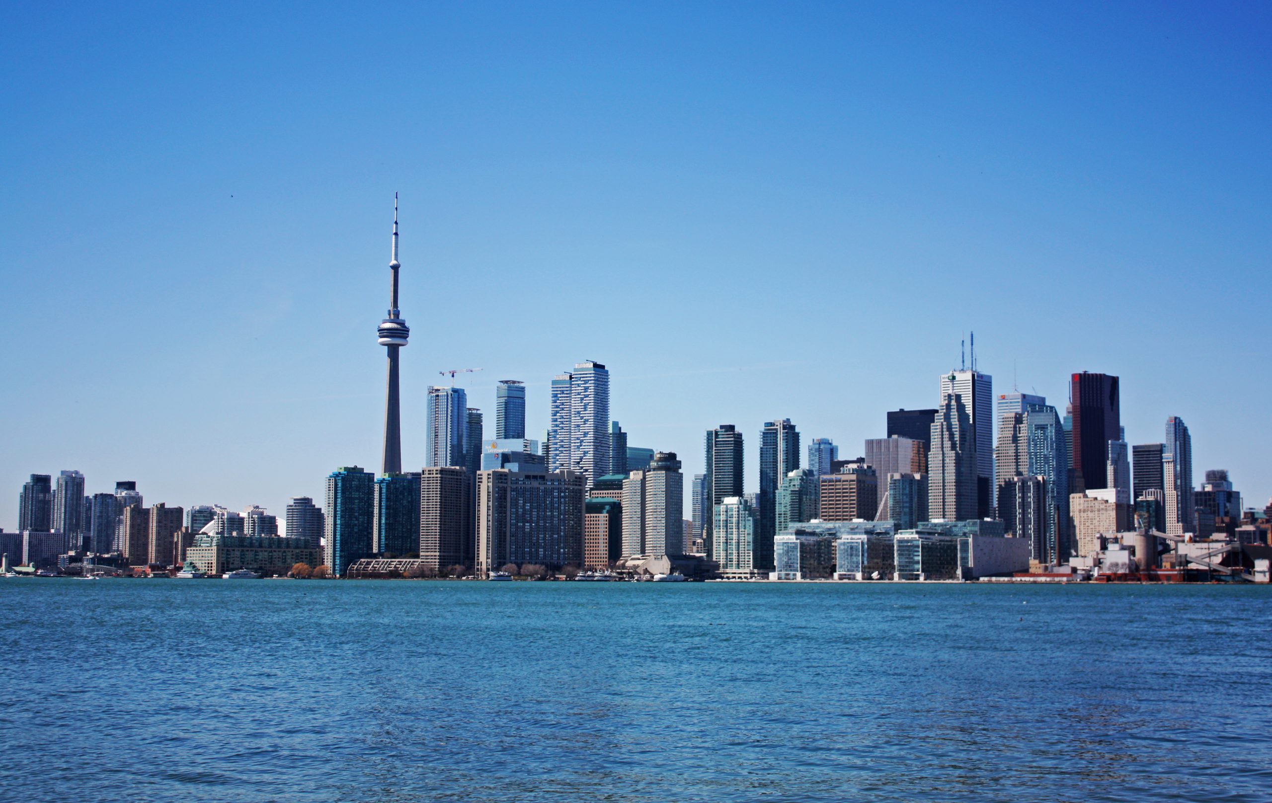 View of downtown Toronto from the water. The CN Tower is surrounded by tall buildings with Lake Ontario in the foreground.