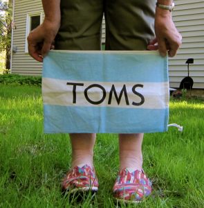 A photo of a person, shown only from the waist down, standing on a lawn wearing colourful TOMS shoes and holding a blue and white stripped TOMS canvas bag above their feet in front of their shins. 