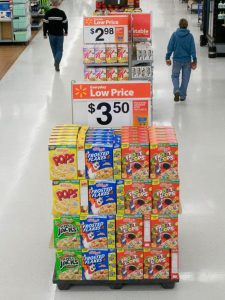 Four columns and four rows of cereal boxes stacked at the end of a supermarket's aisle with a large "low price" sign across the top.