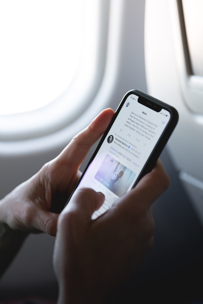Two Hands holding an iphone X displaying a social media page; the background is an airplane window.