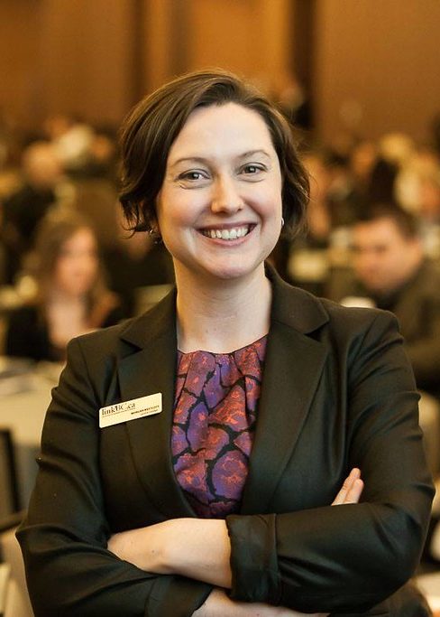 A smiling young woman in a blazer and name tag poses with her arms crossed.