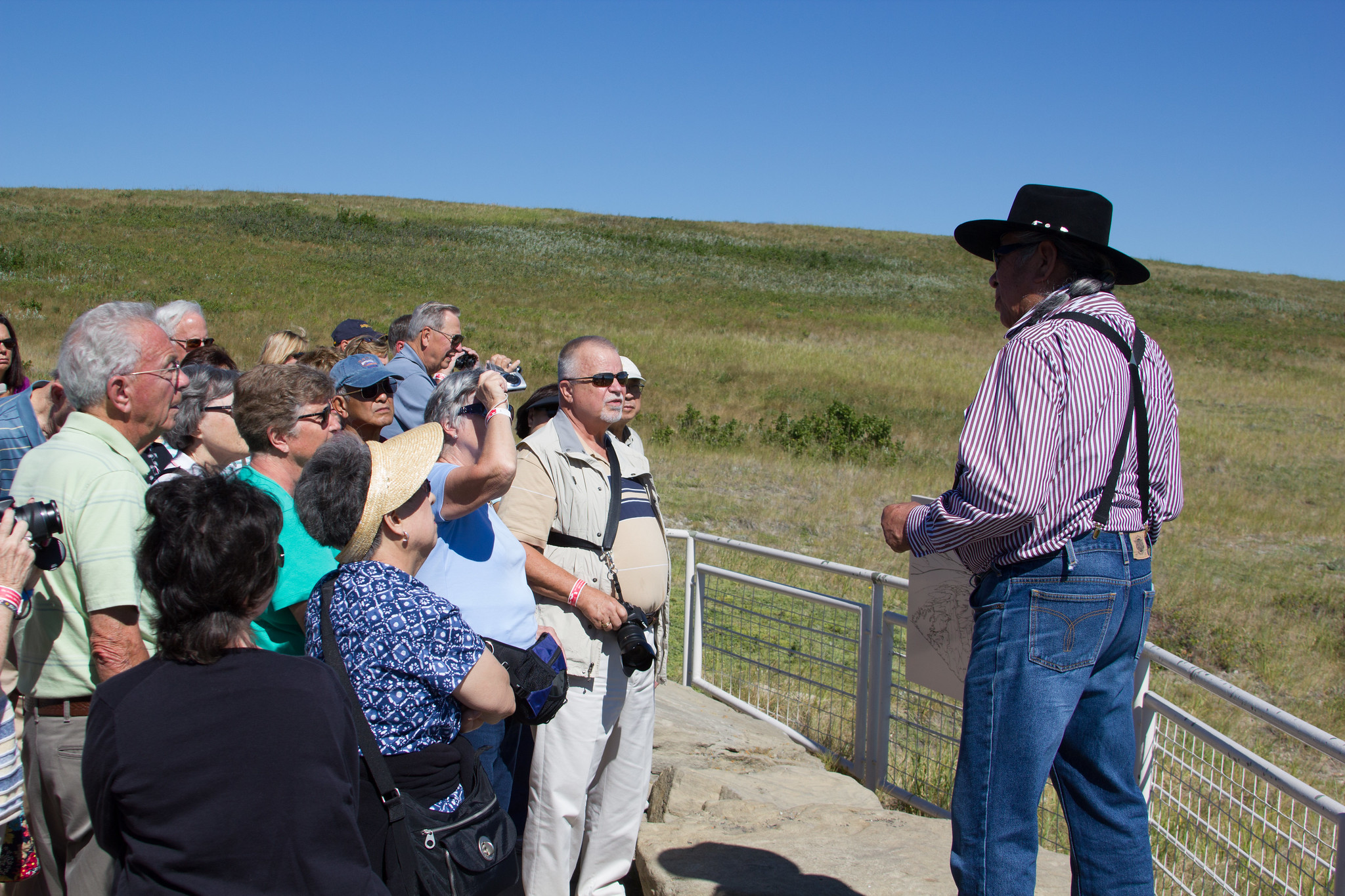 Tourists gather around a man wearing a cowboy hat next to a short metal fence in front of a plain.