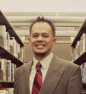 A man in a suit and tie smiles in between bookshelves in a library.
