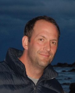 A man in a puffy jacket smiles by the ocean in the evening.
