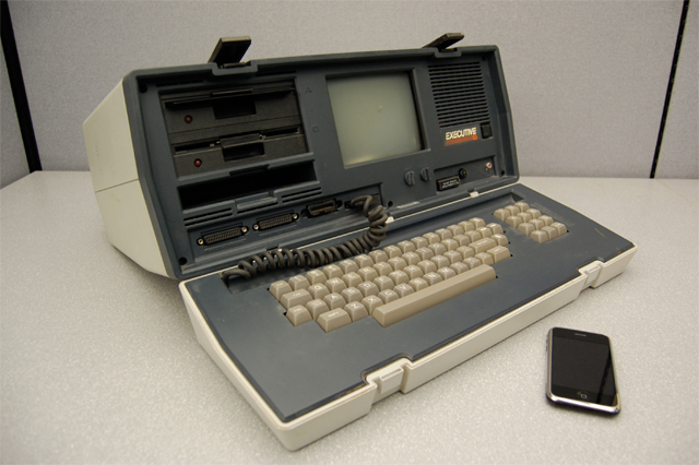 A large, old computer (roughly 2 feet by 1 foot large) with an iphone sitting next to it