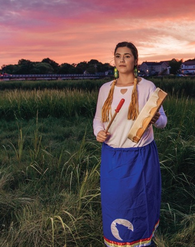A young woman is standing in a field at sunset dressed in traditional Mi’kma’ki clothing holding a drum.