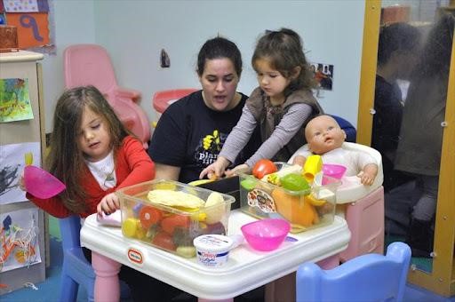 an educator kneeling down beside two children in a dramatic play area. There is a baby doll in a high chair, food and dishes on a child-sized table.