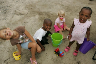four children playing in the sand with pails, shovels and small plastic toy cars