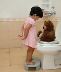 Photo of a child standing on a stool in front of the toilet. Her stuffed bear is sitting on the toilet seat.