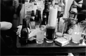 A black and white image of a table of drinks and empty alcohol bottles.