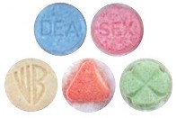 5 different colored MDMA/Ecstacy tablets. There is a blue one with DEA imprinted on it, a pink one with SEX imprinted on it, a cream colored one with WB imprinted on it in the style of the Warner Brothers logo, an orange triangular one, and a green one with an X imprinted on it.