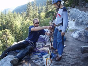 An adult man and a child on a cliff. The man is helping the child adjust their rock-climbing equipment.