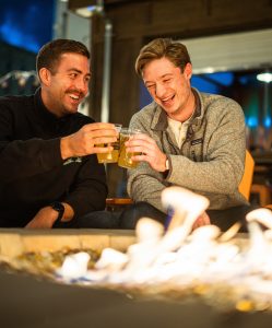 Two smiling men clinking their cups of beer together by a bonfire.
