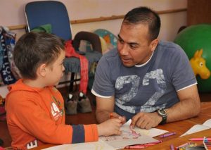 A father interacting with his son who is drawing a picture. He could be portraying the style of teacher-counselor or athletic coach.