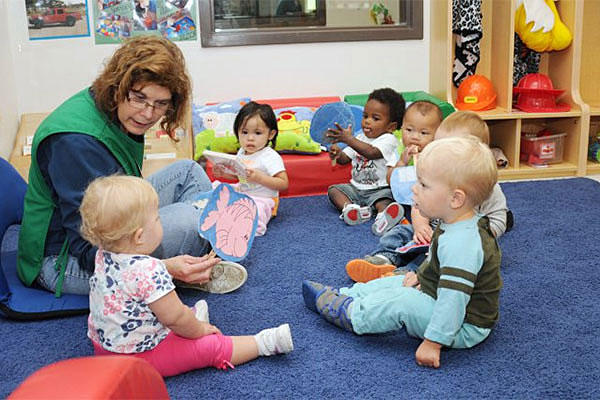 Six toddlers sitting on the rug in a circle looking at a teacher.