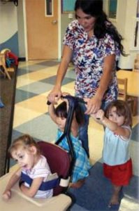 Three children and a teacher, child with cerebral palsy is strapped into a special chair that helps her stay upright and safe