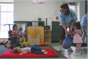 Three children and a teacher in a classroom, one child is lying on a mat.