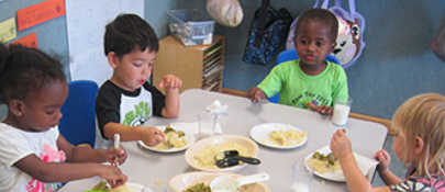 Four young children sitting at the table eating lunch and serving themselves from serving bowls