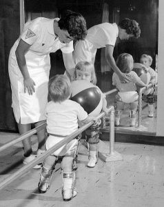 Two young children walking with braces while a nurse looks on.