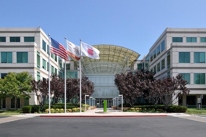 Photograph of Apple Inc.’s headquarter buildings in Cupertino, California. Two squarish buildings with an arched-roof in between. The United States, California and Apple flags are flying in the open-air entrance.