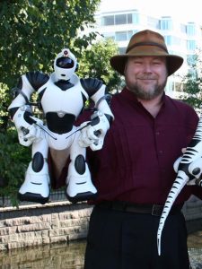 35-45 year old man in a Tilley hat standing by a wall and holding his robot creation (approximately 14 inches tall)