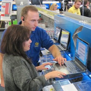A 20-30 year old blonde man assisting a 40-50 year old brunette woman in front of a laptop.