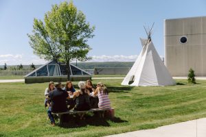gathering of students seated on a picnic table outdoors, while a tipi stands in the backdrop on the NSCC campus.