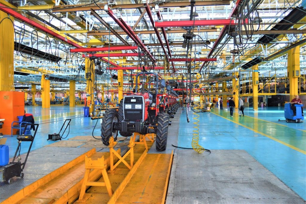 The interior of a factory. A very large space filled with pillars and cords. In the centre a line of tractors.