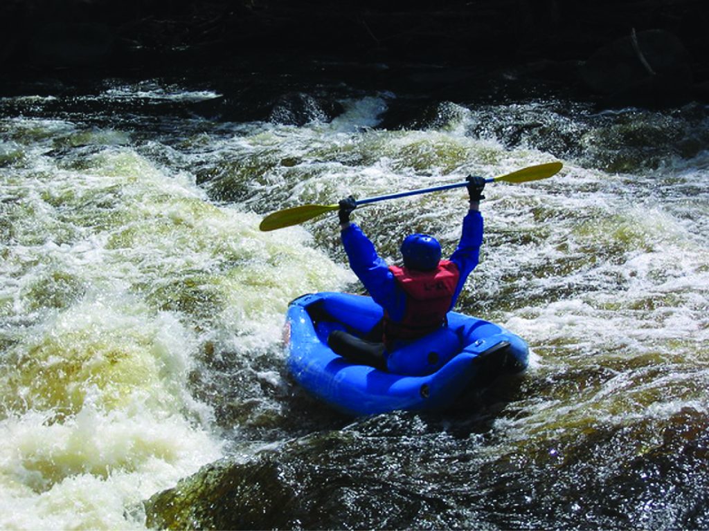 A single person river rafting in a blue inflatable boat, wearing a lifejacket and holding their oar above their head.