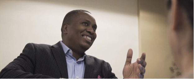 A man wearing a blue shirt and dark coat is smiling and gesturing with his hand in conversation.