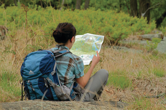 A person sits on the ground outdoors wearing a backpack and a plaid shirt is looking at a paper map.