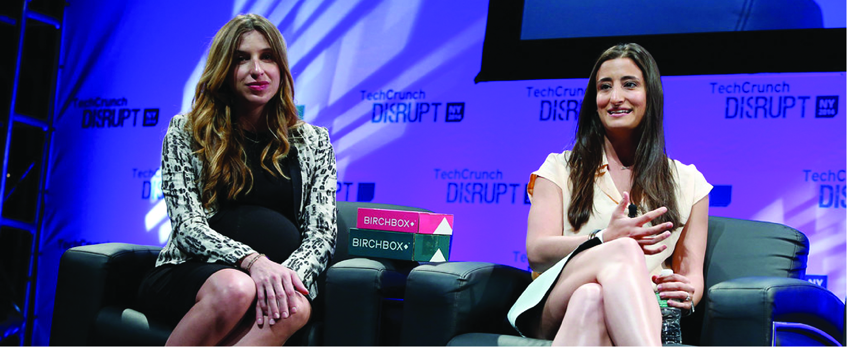 Two women sitting in black chairs in front of a screen.