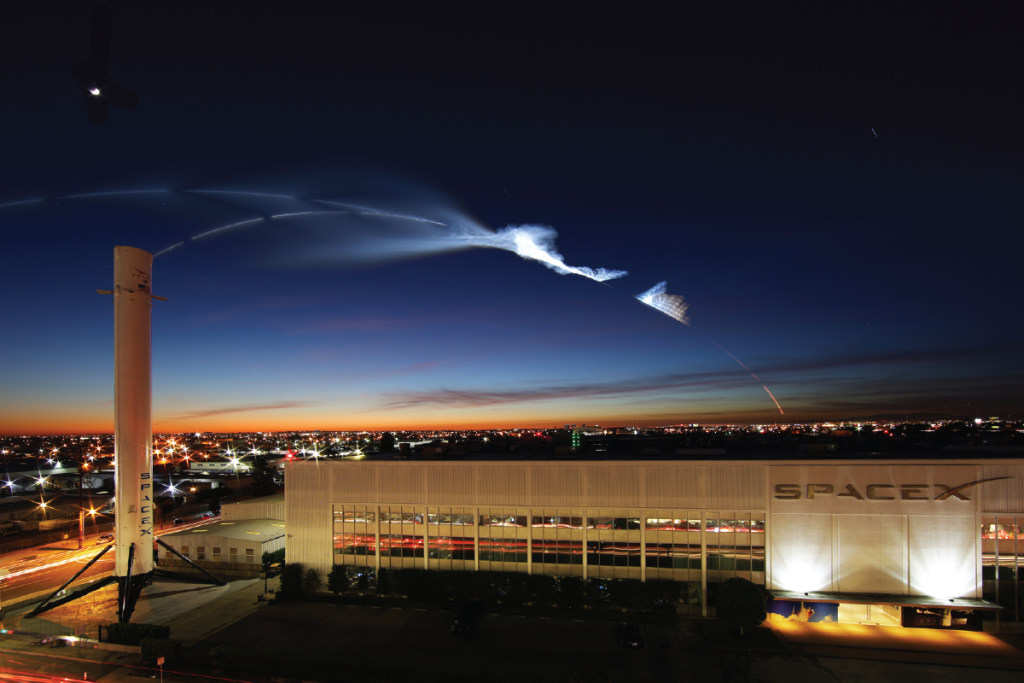 A white industrial building at dusk, with a rocket launch trail in the sky behind it.