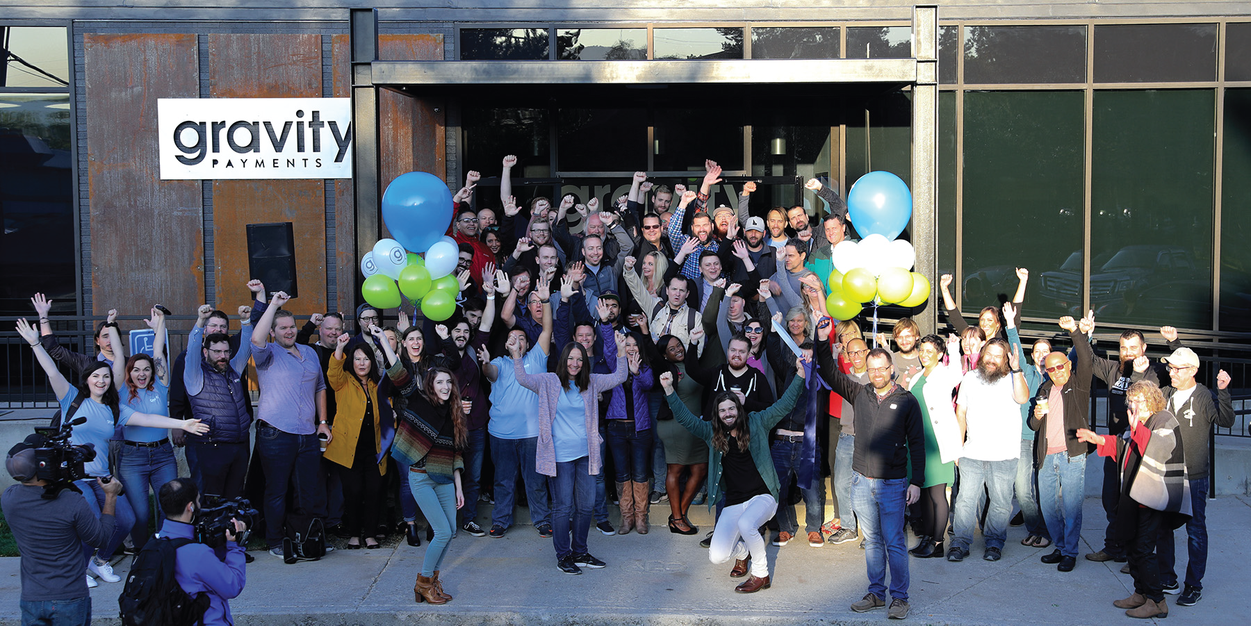 A large group of people stand outside the door of a business, their arms in the air in celebration.