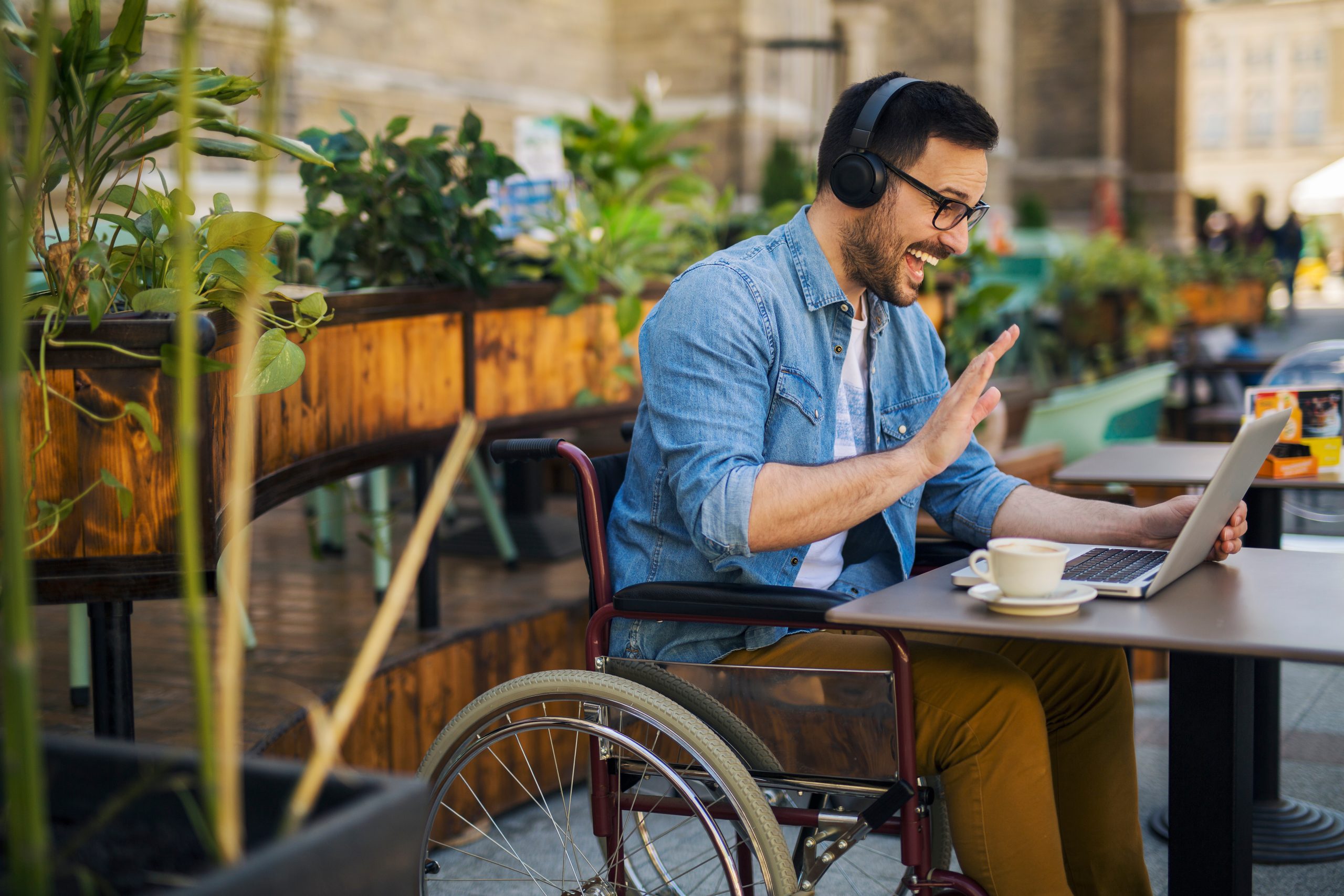 Young man at an outdoor cafe table, wearing glasses, denim shirt and brown pants. Person is talking to someone on a computer and has a headset on.