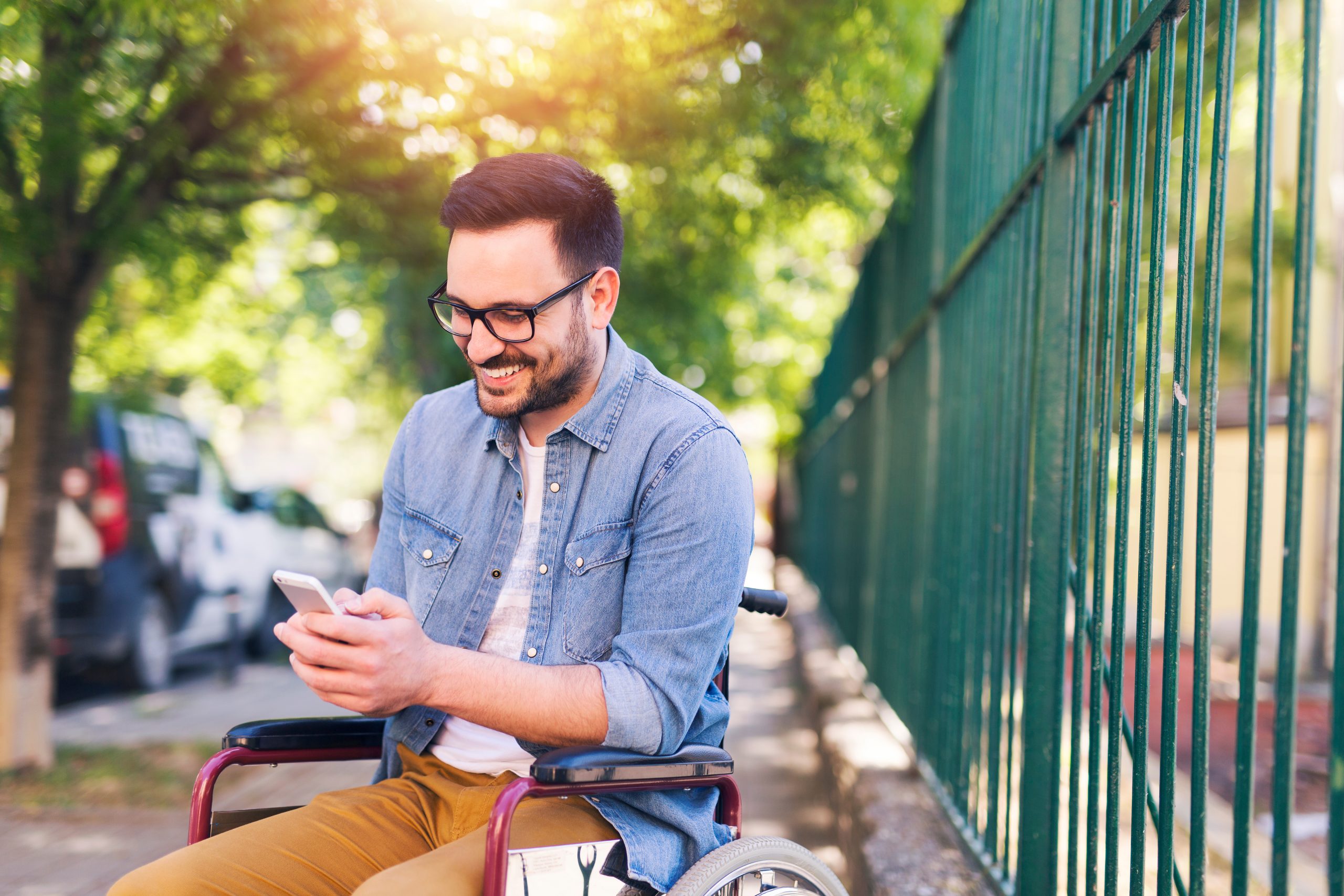 Young man with brown hair, glasses, white tshirt and denim shirt sitting in a wheelchair outside. He is smiling at his phone.