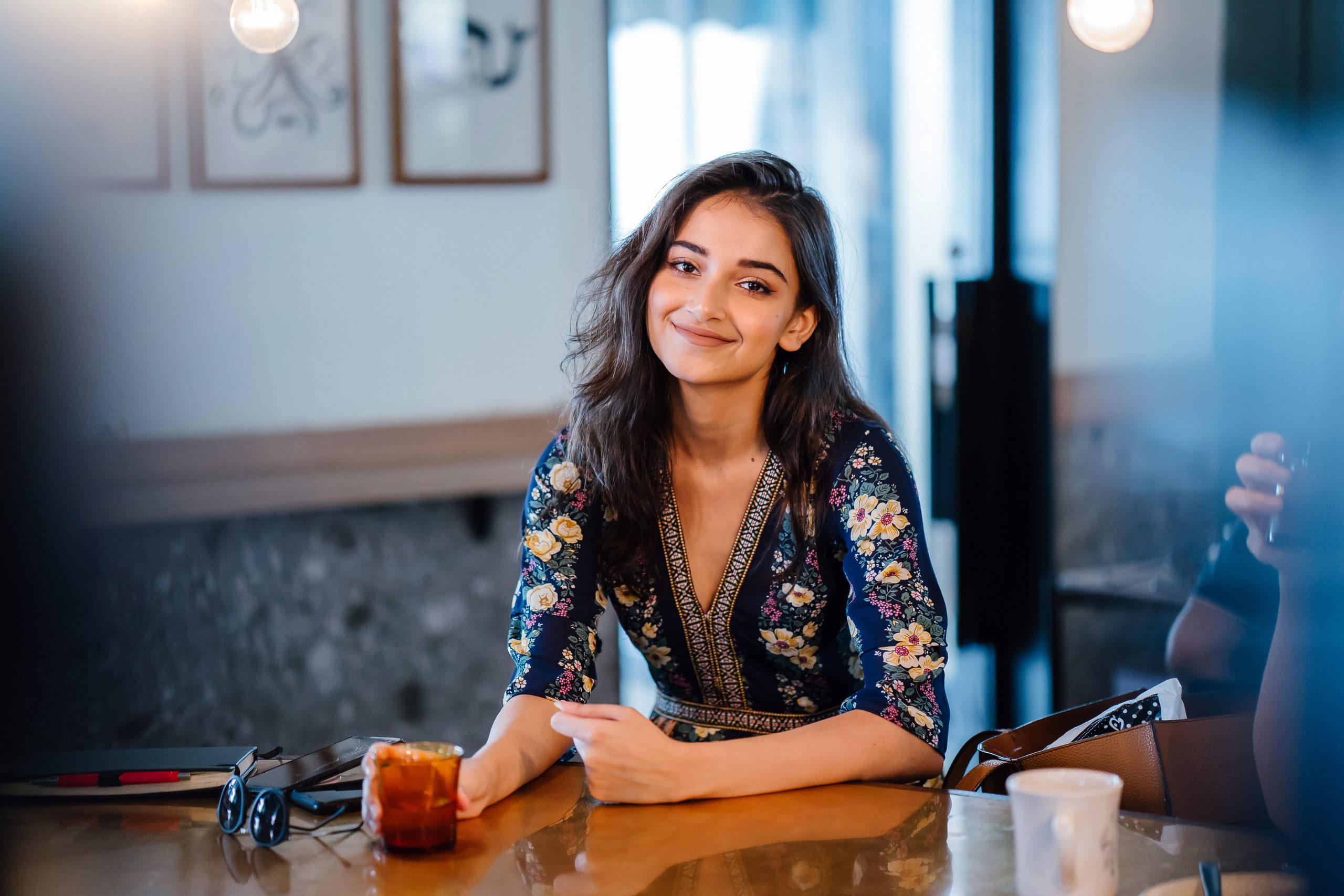 Young woman in a coffee shop sitting at a table. She has a patterned top and shoulder length dress and is smiling at the camera.