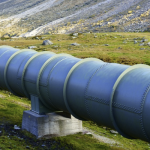 The image shows a series of large, cylindrical pipeline in a grassy field, with mountains visible in the background