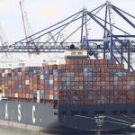 The image shows a large cargo ship with multiple stacks of shipping containers on its deck