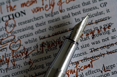 A pen poised on top of typed page that contains many corrections in red ink