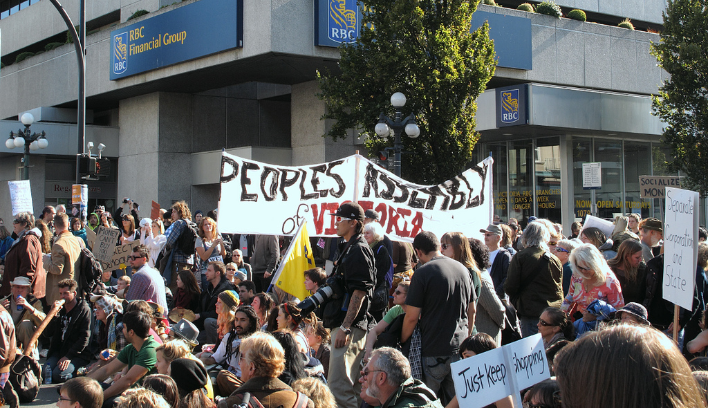 A large crowd of people gathered on the street. One sign says, "Peoples Assembly of Victoria."