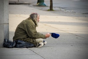 An old man in ratty clothes sits on the corner of the street holding out a hat for money.