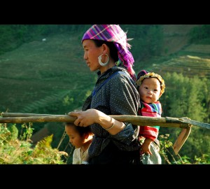 A woman carries tools in one arm with a baby on her back and a young girl by her side.