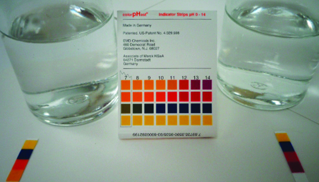 This photo shows two glass containers filled with a transparent liquid. In between the containers is a p H strip indicator guide. There are p H strips placed in front of each glass container. The liquid in the container on the left appears to have a p H of 10 or 11. The liquid in the container on the right appears to have a p H of about 13 or 14.