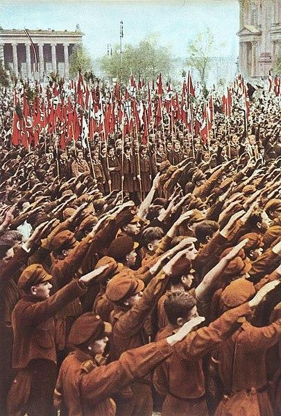 Young boys in the Hitler Youth throwing the Nazi salute with arms upstretched, with girls in the League of German Girls holding Nazi flags.