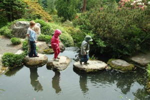 Four children walking on stepping stones to get accross a small pond.
