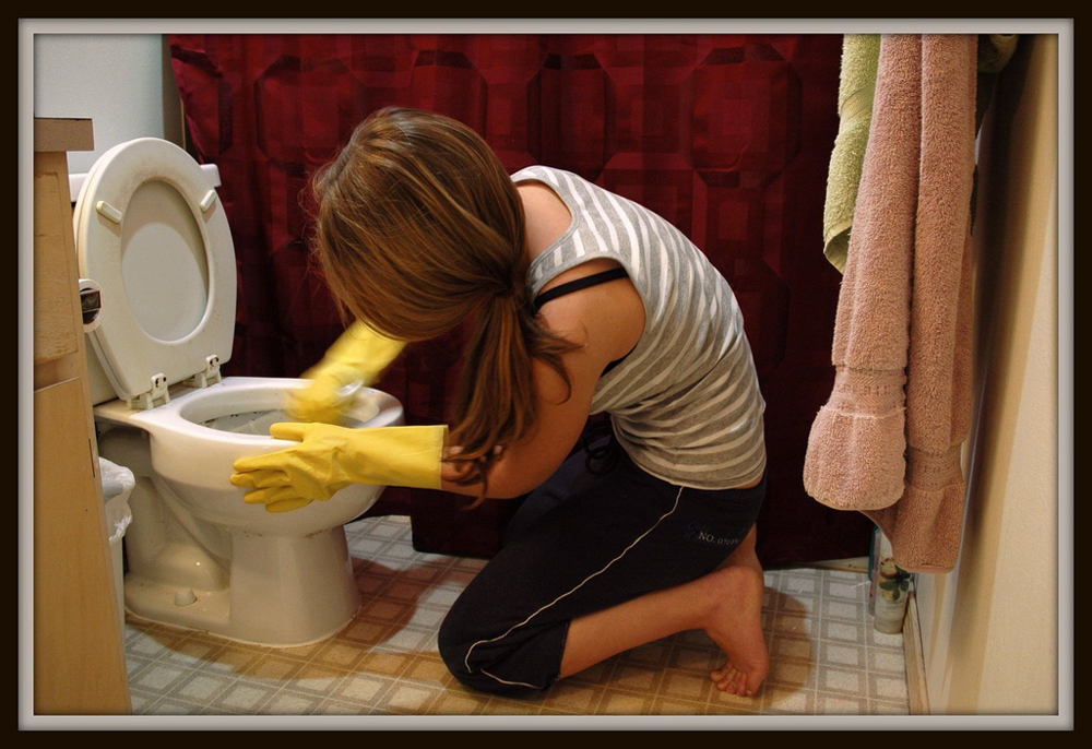 A woman kneeling on bathroom floor scrubbing a toilet.