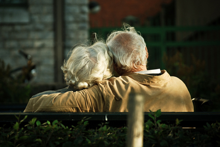An old couple sitting on a park bench leaning into each other.