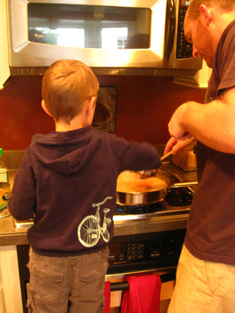 A young boy stands at a stove beside a man helping stir a pot.