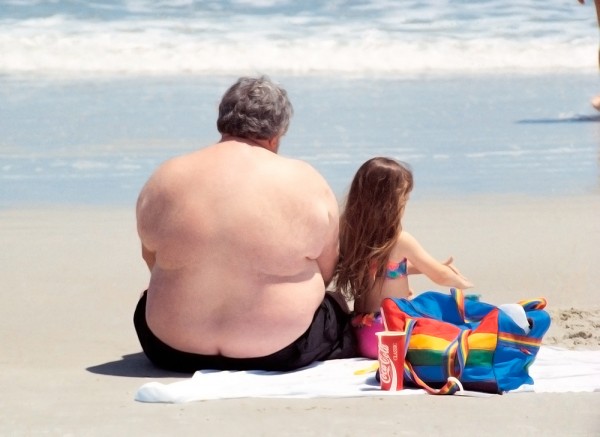 An overweight older man sits on a beach in his swimsuit beside a young girl playing in the sand.